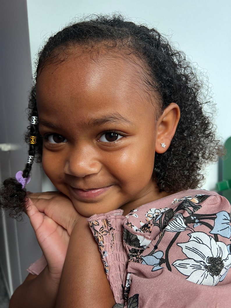 May include: A young girl with curly dark brown hair wearing a pink floral dress and small silver earrings. She is smiling and looking at the camera.