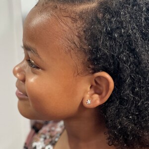 May include: A young girl with curly black hair wearing a small flower-shaped earring in her left ear.