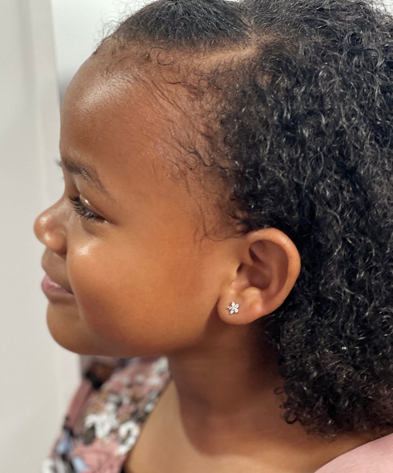 May include: A young girl with curly black hair wearing a small flower-shaped earring in her left ear.