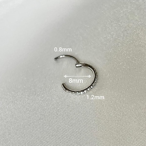 May include: A silver hoop earring with a row of clear cubic zirconia stones. The earring is 8mm in diameter and 1.2mm wide.