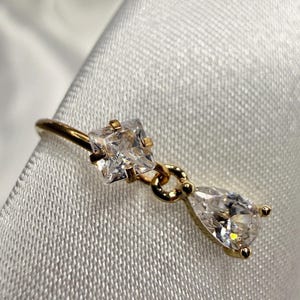 May include: A gold ring featuring two clear, sparkling cubic zirconia stones. One stone is square-cut, and the other is pear-shaped, suspended from a delicate gold band. The ring is set against a white, textured background.