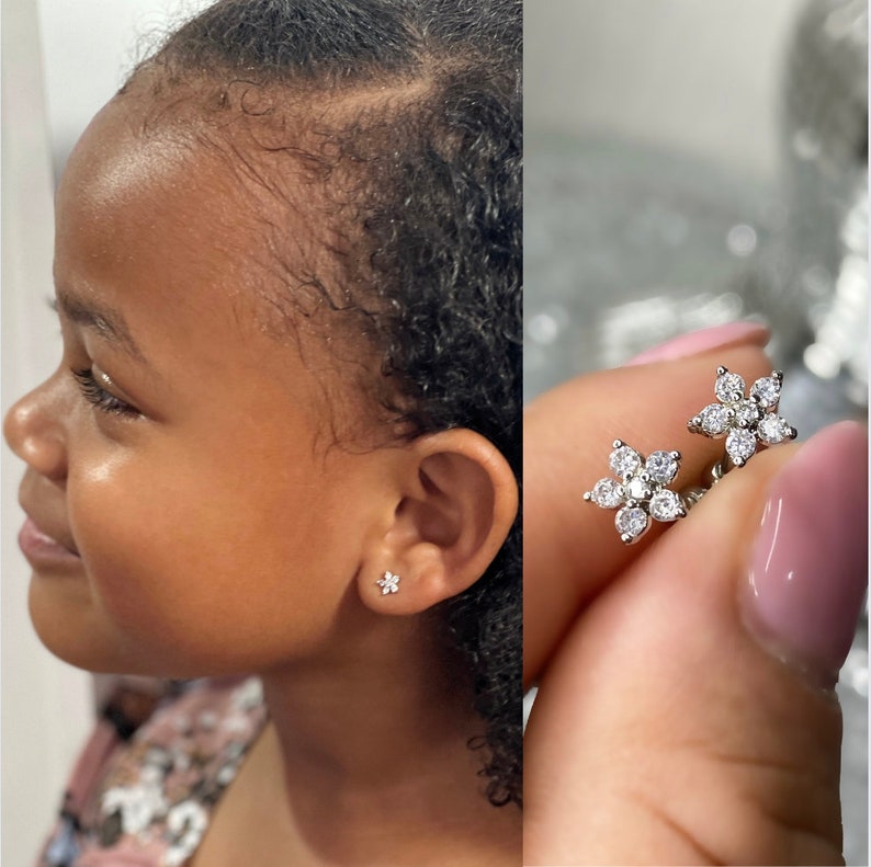 May include: A pair of silver flower-shaped earrings with clear cubic zirconia stones. The earrings are shown on a person's ear and in a close-up view.