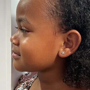 May include: A young girl with curly black hair wearing a pink and floral patterned shirt. She is wearing a small flower-shaped earring in her left ear.