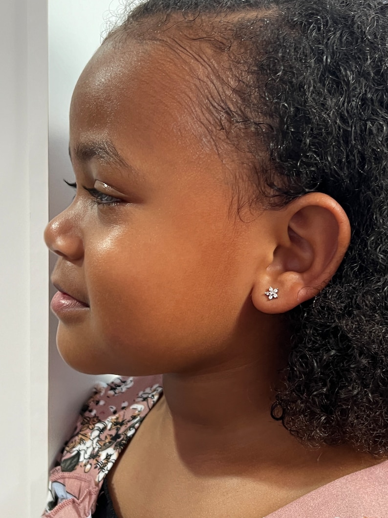 May include: A young girl with curly black hair wearing a pink and floral patterned shirt. She is wearing a small flower-shaped earring in her left ear.