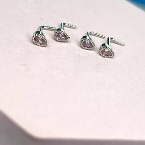 May include: Four silver-toned stud earrings with a pear-shaped, pink gemstone center. The earrings are set with small, clear stones around the pink gem. The earrings are displayed on a light pink surface.
