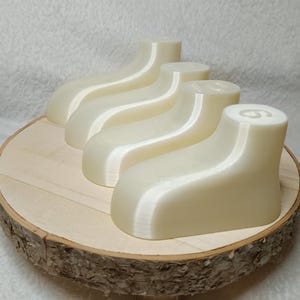 Könnte beinhalten: Ein Set aus vier cremeweißen, glatten und geschwungenen Kunststoffformen, die stilisierten Füßen oder Schuhleisten ähneln. Jedes Teil hat eine abgerundete Ferse und eine zulaufende Spitze, wobei die Zahl sechs auf der Oberseite eingeprägt ist.
