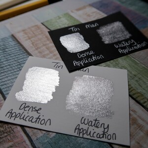 May include: Two swatches of metallic silver paint on a white card. The swatches show the paint applied in a dense and watery application. The card is labeled "Tin Man" and the swatches are labeled "Dense Application" and "Watery Application".