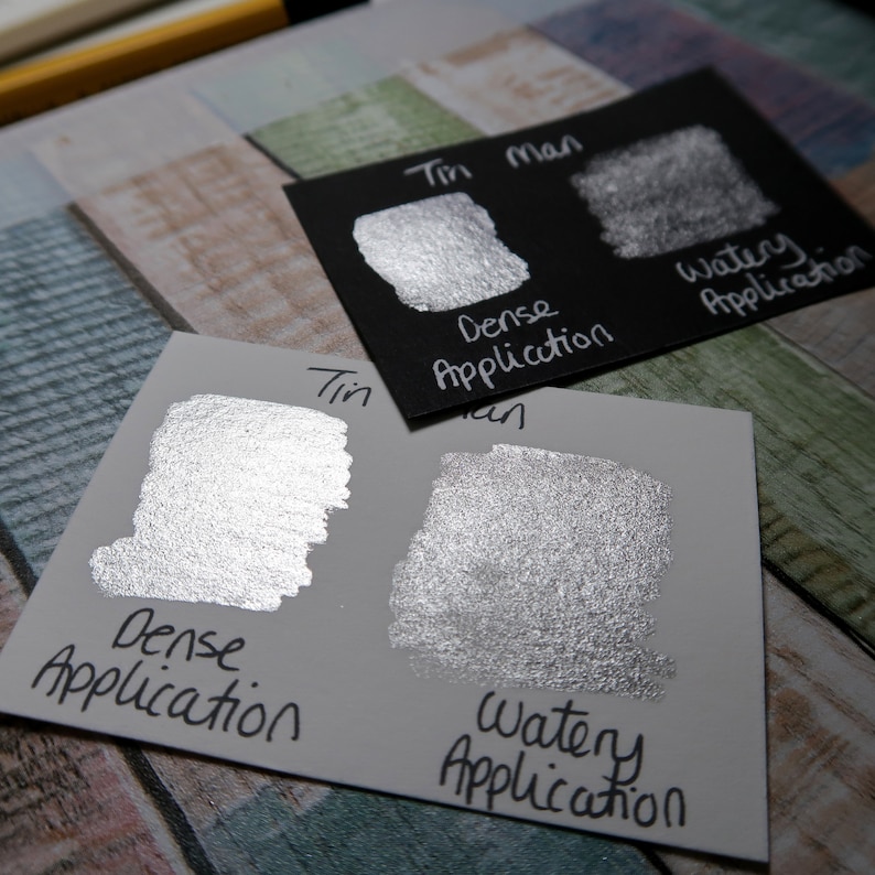 May include: Two swatches of metallic silver paint on a white card. The swatches show the paint applied in a dense and watery application. The card is labeled "Tin Man" and the swatches are labeled "Dense Application" and "Watery Application".