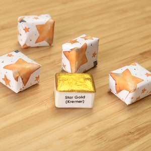 Może przedstawiać: Kolekcja farb akwarelowych, w tym złota farba metaliczna z napisem "Star Gold (Kremer)". Farby znajdują się w małych, kwadratowych pojemnikach. Niekt&oacute;re są owinięte papierem ze wzorami pomarańczowych gwiazd.