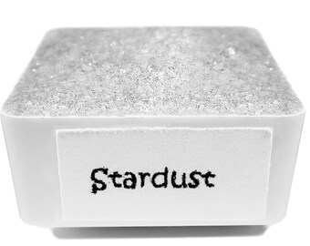 May include: A small white square container filled with a sparkly silver substance. The container has a white label with the word "Stardust" printed on it.