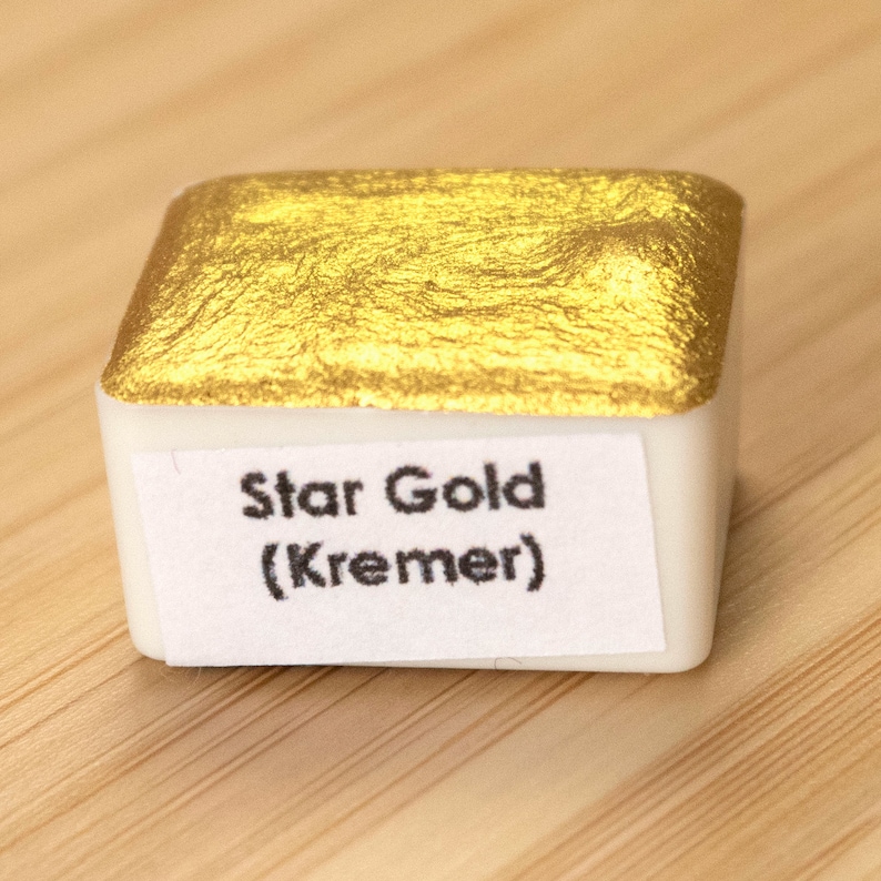 Może przedstawiać: Kwadratowa patelnia z farbą akwarelową o metalicznej, złotej powierzchni. Etykieta głosi "Star Gold (Kremer)" czarnym tekstem. Farba znajduje się w białym plastikowym pojemniku, na jasnobrązowej drewnianej powierzchni.