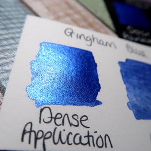May include: A close-up of a blue ink sample with a metallic sheen. The ink is labeled "Gingham Blue" and "Dense Application".