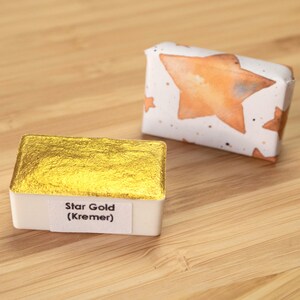 Może przedstawiać: Prostokątny pojemnik z farbą akwarelową w kolorze złotym, oznaczony jako "Star Gold (Kremer)". Obok niego mały, zapakowany pakunek ze wzorem pomarańczowych i białych gwiazd. Na drewnianej powierzchni.