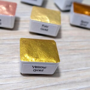 May include: A small square pan of yellow gold watercolor paint. The pan is labeled "Yellow Gold".