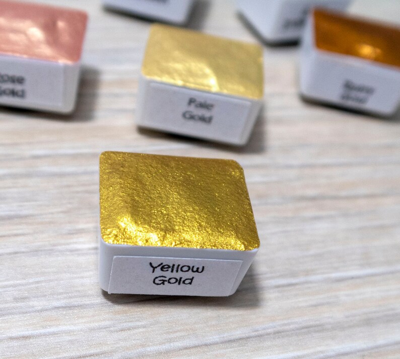 May include: A small square pan of yellow gold watercolor paint. The pan is labeled "Yellow Gold".
