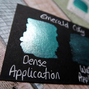 May include: A close-up of a teal green metallic paint swatch on a black card. The card has the words "Emerald City" and "Dense Application" written in white.