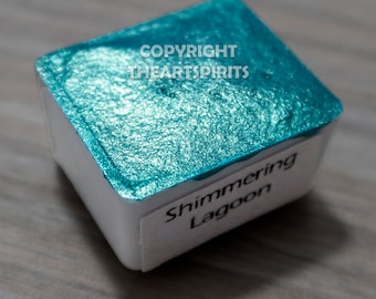May include: A square watercolor paint pan filled with shimmering teal paint. The paint is labeled "Shimmering Lagoon" on a white label. The top of the pan has the text "COPYRIGHT THEARTSPIRITS".