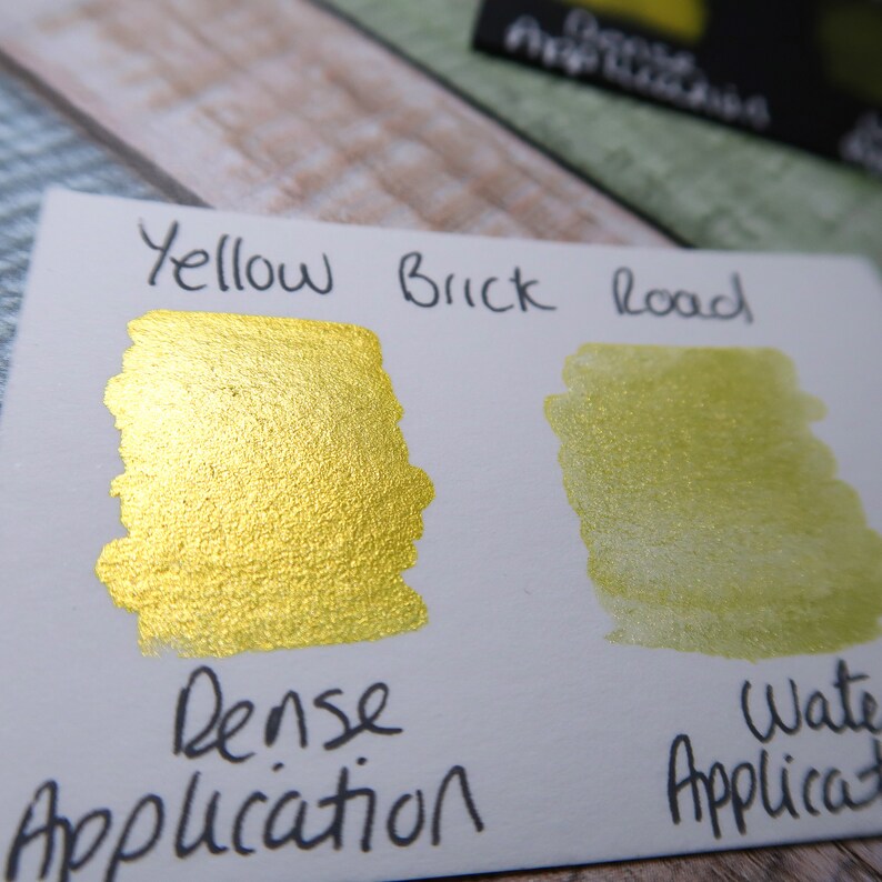May include: A white card with the words "Yellow Brick Road" written in black ink. Two swatches of yellow paint are shown, one with a dense application and the other with a water application.