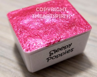 May include: A square watercolor paint pan with a vibrant, shimmering pink surface. The white label on the side reads "Sleepy Poppies" in a handwritten font. The top of the pan has the text "COPYRIGHT THEARTSPIRITS".