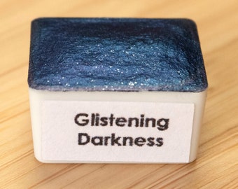May include: A small, square, white pan of watercolor paint with a blue, glittery top. The label reads "Glistening Darkness".