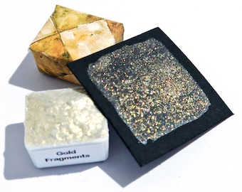May include: A small white square container with the text "Gold Fragments" on the label. The container is filled with a glittery gold paint. A sample of the paint is shown on a black card.