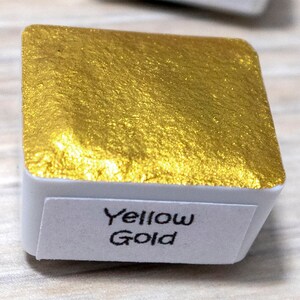 May include: A small, square, white container with a gold metallic paint inside. The container has a label that reads "Yellow Gold".