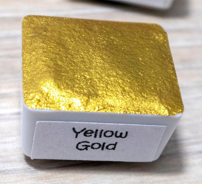 May include: A small, square, white container with a gold metallic paint inside. The container has a label that reads "Yellow Gold".
