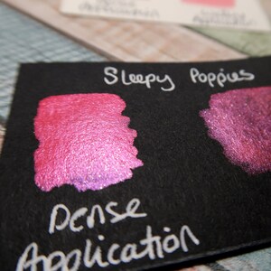 May include: A black card with the words "Sleepy Poppies" written in white. Two swatches of pink shimmery ink are shown on the card. The text "Dense Application" is written in white below the swatches.