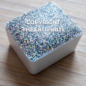 以下が含まれることがあります： 虹色の銀色のグリッターで満たされた白い正方形の容器。グリッターの上に「COPYRIGHT THEARTSPIRITS」というテキストが印刷されています。