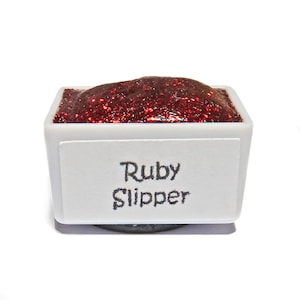 May include: A small white pan containing red glitter paint. The pan has a label that reads "Ruby Slipper".