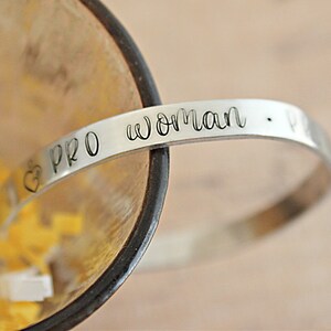 May include: A silver-colored bracelet with the words "PRO woman" engraved on it. A heart symbol is next to the word "PRO". The bracelet is curved and sits inside a wooden bowl with yellow and white confetti.