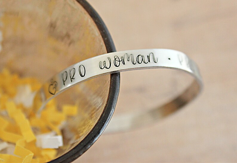 May include: A silver-colored bracelet with the words "PRO woman" engraved on it. A heart symbol is next to the word "PRO". The bracelet is curved and sits inside a wooden bowl with yellow and white confetti.