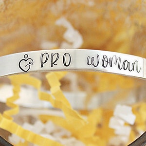 May include: A silver-colored cuff bracelet with the words "PRO woman" engraved on it. The bracelet features a heart-shaped design with a stylized figure inside. The bracelet is displayed inside a clear glass container with yellow paper filler.