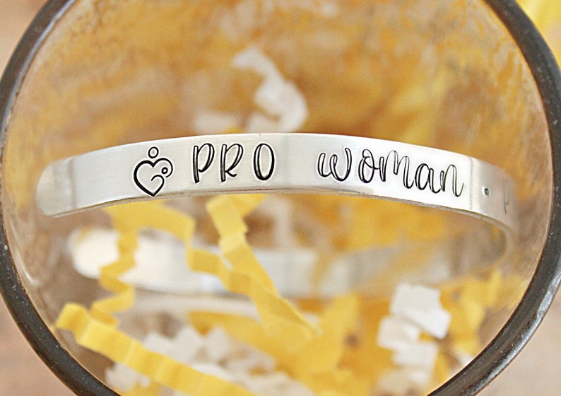 May include: A silver-colored cuff bracelet with the words "PRO woman" engraved on it. The bracelet features a heart-shaped design with a stylized figure inside. The bracelet is displayed inside a clear glass container with yellow paper filler.