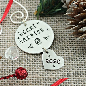 May include: Silver Christmas ornament with the words "heart warrior" and a heart graphic. A smaller heart charm hangs below with the year "2020" inscribed.