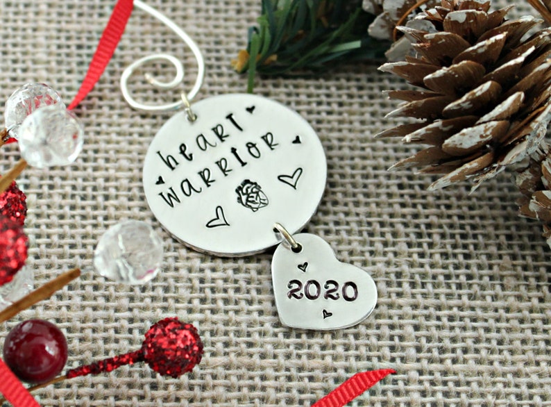 May include: Silver Christmas ornament with the words "heart warrior" and a heart graphic. A smaller heart charm hangs below with the year "2020" inscribed.