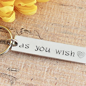 May include: A silver rectangular keychain with the phrase "as you wish" and a heart-shaped Celtic knot design. The keychain is attached to a silver ring. Yellow flowers are in the background.
