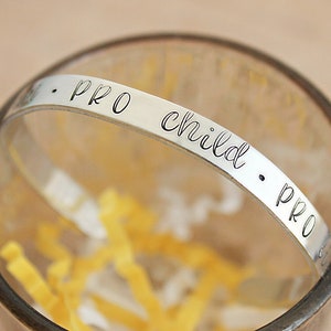 May include: A silver-colored bracelet with the words "PRO child PRO" engraved in a delicate, cursive font. The bracelet is inside a clear glass, with yellow paper filling the bottom. The bracelet is a simple, elegant piece of jewelry.