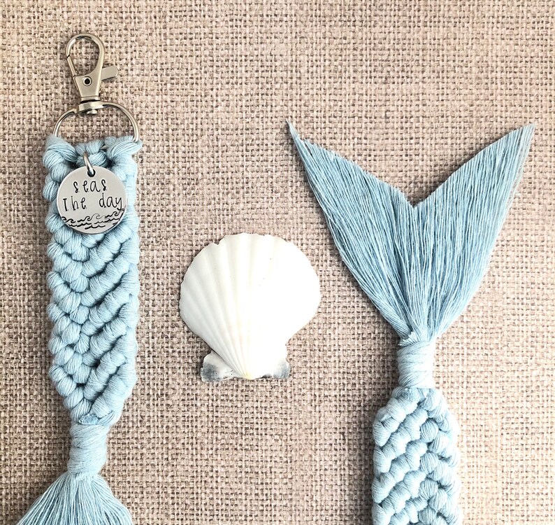 May include: A light blue macrame keychain with a silver charm that says "Seas the day". The keychain is next to a light blue macrame mermaid tail.