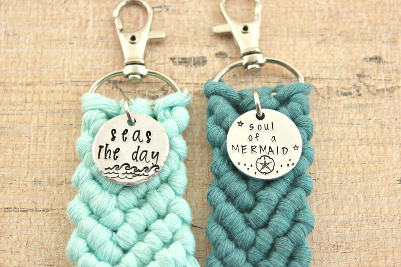 May include: Two crocheted keychains with silver charms. The keychain on the left is light blue and has a charm that says "Seas the day" with a wave design. The keychain on the right is teal and has a charm that says "Soul of a Mermaid" with a starfish design.