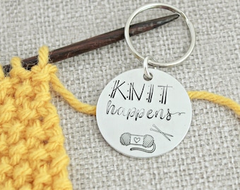 May include: A silver keychain with the text "Knit Happens" and a small illustration of yarn and knitting needles. The keychain is attached to a brown knitting needle and yellow yarn.