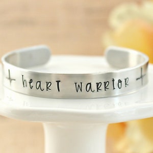 May include: Silver cuff bracelet with the words "heart warrior" and a cross on each side.
