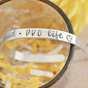 May include: A silver-colored cuff bracelet with the words "child &bull; PRO life" engraved on it. The bracelet also features a heart with a baby inside. The bracelet is resting inside a clear glass.
