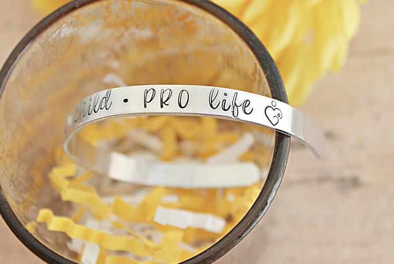May include: A silver-colored cuff bracelet with the words "child &bull; PRO life" engraved on it. The bracelet also features a heart with a baby inside. The bracelet is resting inside a clear glass.