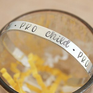 May include: A silver-colored bracelet with the words "PRO child PRO" engraved in a delicate, cursive font. The bracelet is set inside a clear glass, with yellow paper strips at the bottom. The bracelet is a piece of jewelry.