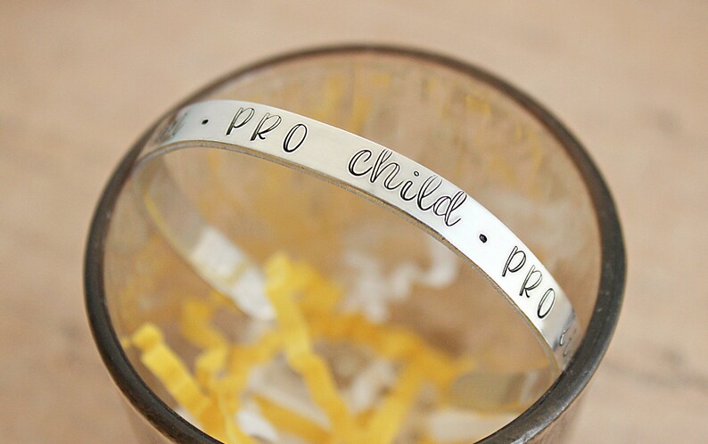 May include: A silver-colored bracelet with the words "PRO child PRO" engraved in a delicate, cursive font. The bracelet is set inside a clear glass, with yellow paper strips at the bottom. The bracelet is a piece of jewelry.