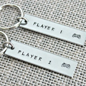 May include: Two silver keychains with key rings. One keychain has the text "PLAYER 1" and a small image of a video game controller. The other keychain has the text "PLAYER 2" and a small image of a video game controller.
