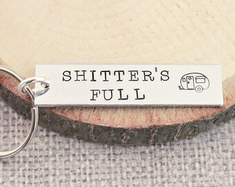 Könnte beinhalten: Silberner Schlüsselanhänger mit einem kleinen Wohnwagen-Symbol und dem Text "Shitter's Full" auf einer Holzoberfläche.