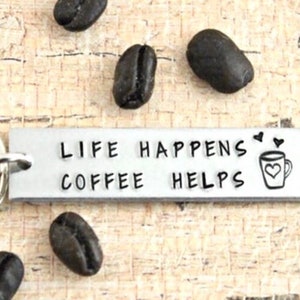 May include: Silver keychain with a metal tag that reads "Life Happens Coffee Helps" with a small coffee cup icon and two hearts. The keychain is surrounded by coffee beans.