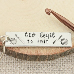 May include: Silver keychain with a rectangular tag that reads "too legit to knit". The tag has a small image of a yarn ball and crochet hook on each side. A brown crochet hook is lying on a wooden surface next to the keychain.